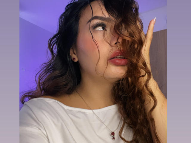 GalaMalkova with wavy brown hair, red lips, white shirt, hand in hair, looking sideways, blue-purple light, small earrings, necklace with red heart, slightly flushed cheeks.