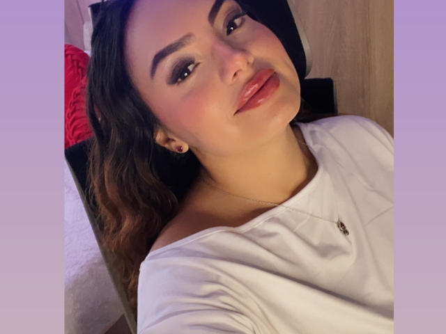 GalaMalkova with shiny pink lips, dark eyebrows, and wavy brown hair, wearing a white shirt, smiling at the camera. She has a nose ring and a necklace.