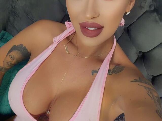 NicolSNaya with tan skin, full lips, pink halter top showing big tits, tattoos on arms, gold necklace, nose ring, and earrings, lying on gray cushioned surface.