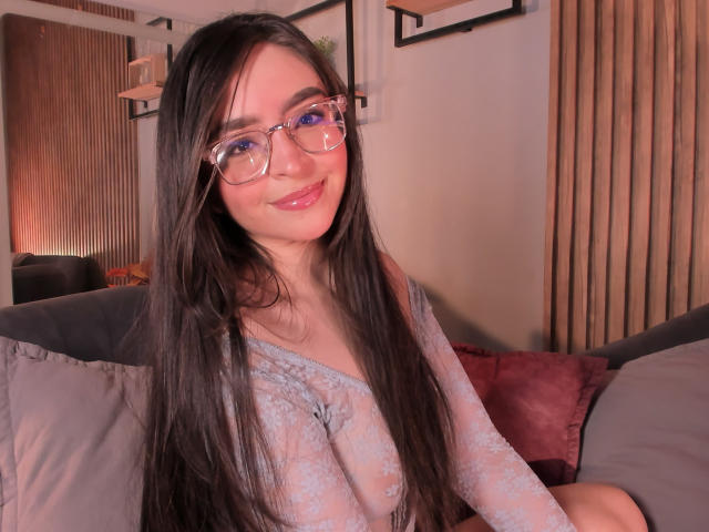 Eva McAllister mit langem, schwarzem Haar, blauen Augen, einer Brille und einem Lächeln, trägt ein durchsichtiges, blaues Oberteil, das kleine Brüste zeigt, und sitzt auf einem Sofa mit roten und grauen Kissen..