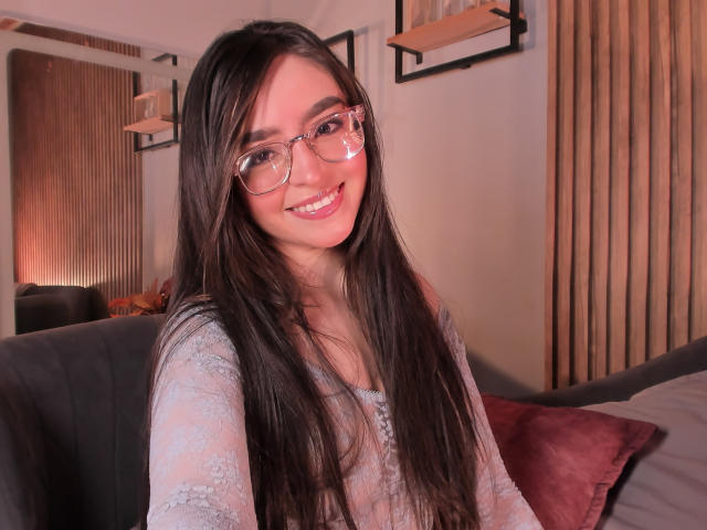 EvaMcAllister with long dark hair, glasses, smiling, wearing a sheer lace top, showing her tits, sitting on a couch, pink pillow in background.