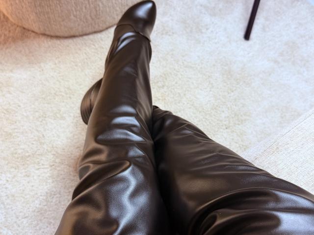 DeliciousChrissy's shiny black leather boots with pointed toes are crossed at the ankle on a beige carpet. The boots have a smooth, slightly wrinkled texture.
