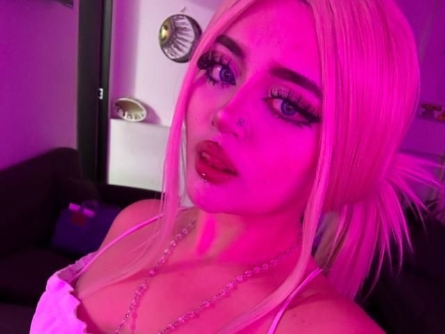 Lyanekkoo with pink hair, heavy eyeliner, and long lashes, has pouty lips and a nose ring. She's wearing a white top with a pearl necklace, under pink lighting.