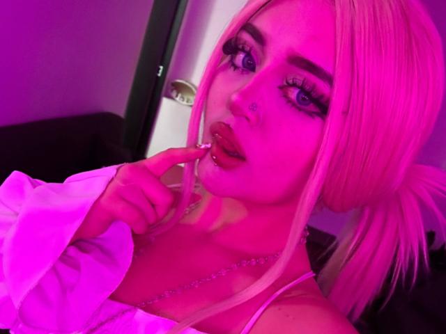 Lyanekkoo with pink hair, blue eyes, and long eyelashes, biting her finger, wearing a white top, pink lighting, nose ring, and necklace, in a selfie.
