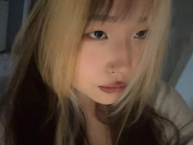 JungMcgranor has blonde and brown hair, a nose ring, and pink lipstick. Her blue eyes are slightly down, with a neutral expression. Her lips are slightly parted.