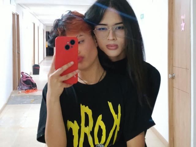 KvsKael with red and black hair, pouting lips, black t-shirt with yellow text, holding red phone, next to KvsKael with long black hair, glasses, and black t-shirt.