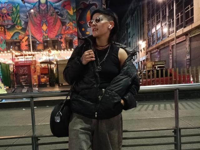 DannyRouds è in una scena notturna di città, indossa una giacca imbottita nera, pantaloni grigi e una borsa nera. Ha un seno piccolo, occhiali e una collana, con un murale che raffigura una città..