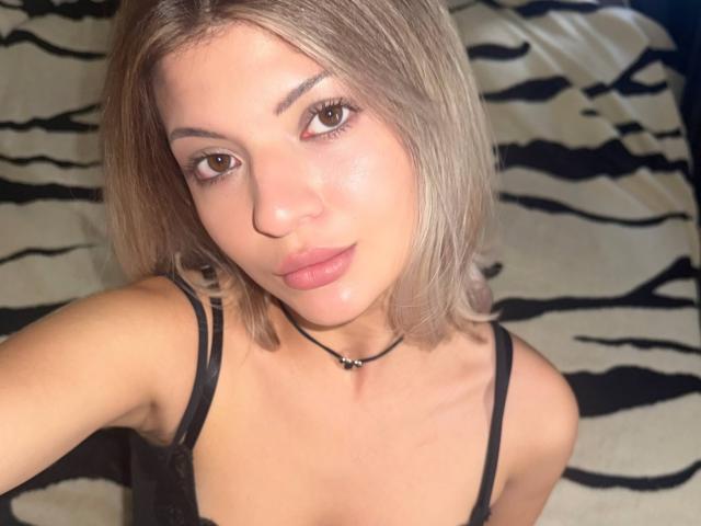 ElianaSemaan has light brown skin, straight blonde hair, brown eyes, and full pink lips. She wears a black choker and a black tank top, showing small tits.