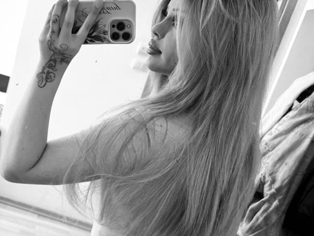 Black-and-white selfie of IvyMirrage with long, straight hair, showing a tattoo on her right wrist, holding a phone, looking to the side, in a mirror.