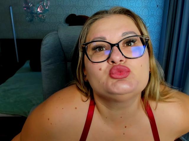 RadaRori with puffy red lips, glasses, blonde hair, red halter top, and a few moles on her face, making a kissy face in a bedroom setting.