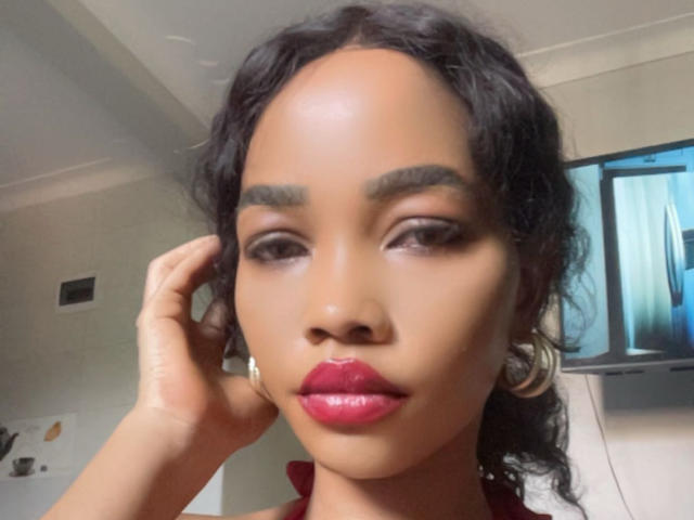 Wizah has shiny brown skin, thick eyebrows, dark wavy hair, and glossy red lips. She touches her head with a hand, looking slightly to the side.