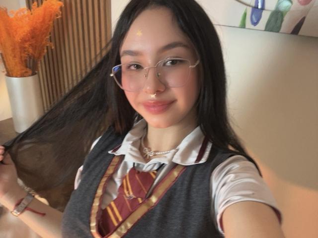 Daphne Valucci, con gli occhiali, il piercing al naso e i lunghi capelli neri, che si tiene i capelli, indossa un'uniforme scolastica grigia con una cravatta a righe rosse e gialle, sorridendo, con un piccolo punto giallo sulla fronte..