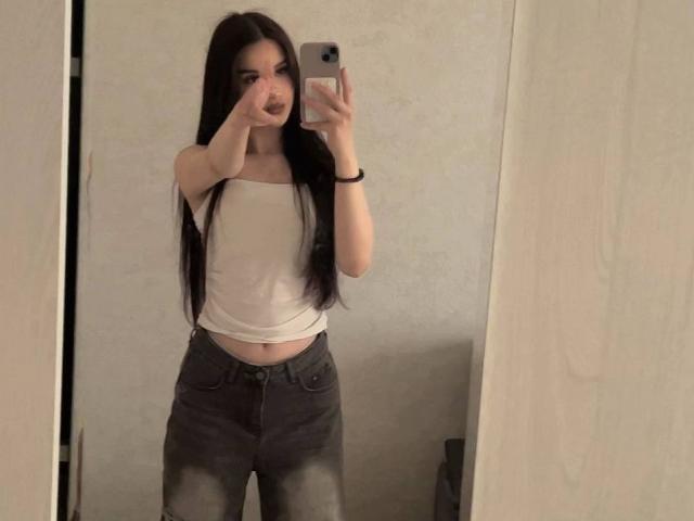 AlixMouton with long dark hair, white strapless top, and gray distressed jeans, taking a mirror selfie with a finger to her mouth, showing her small tits and flat ass.