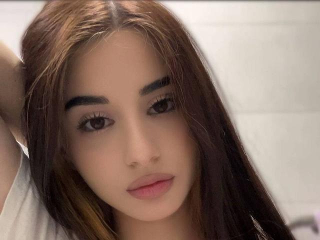 Das Gesicht von AlixMouton, mit ihren langen, braunen Haaren, leicht geöffneten Lippen und braunen Augen, blickt nach vorne und trägt ein kaum merkliches Lächeln. Ihre Hand befindet sich hinter ihrem Kopf, wobei ihre Brüste zur Geltung kommen..
