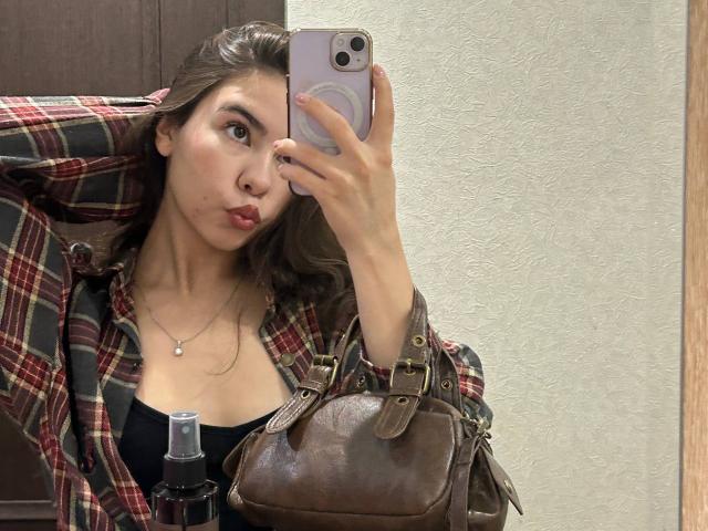 AnneKofron taking a mirror selfie with pouty lips, wearing a plaid shirt over a black top, holding a brown leather bag and a bottle, with long brown hair.