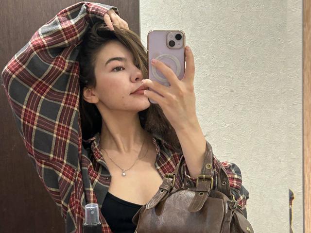 AnneKofron with a plaid shirt, black top, brown bag, and necklace, taking a mirror selfie, sucking her thumb, holding a phone, and looking at the camera.