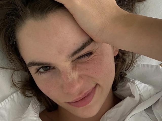 Le visage de PeacHub69, avec un œil fermé, souriant légèrement, une main sur le front, montrant des taches de rousseur, des cheveux bruns ondulés, et portant une chemise blanche, ayant l'air détendu et mignon..
