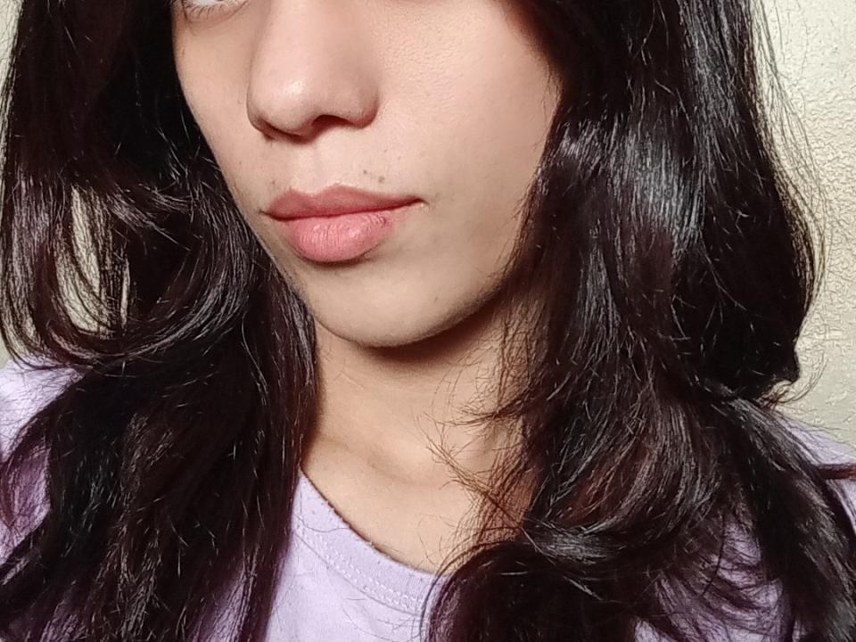 KamiGioLove's face, showing light brown skin, dark wavy hair, pinkish lips, slight pimple on her upper lip, wearing a purple shirt, soft lighting, slight smile.