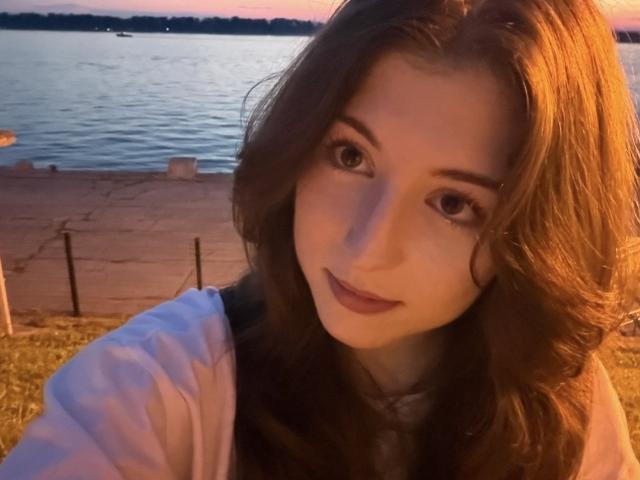 SheilaCiers with wavy brown hair, soft pink lips, and brown eyes, wearing a white shirt, in a sunset by the water. Her tits are small, and she's cute.
