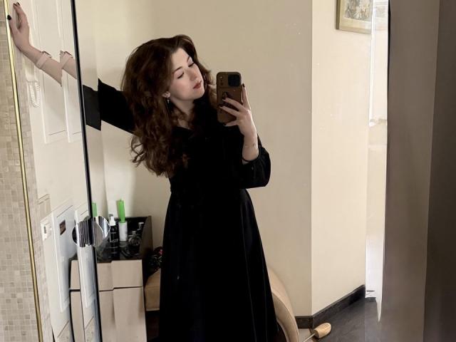 SheilaCiers with curly brown hair, wearing a black dress, taking a mirror selfie, her right arm raised, holding a phone with a gold case, looking slightly to the side.