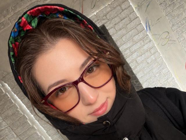 SheilaCiers with glasses, brown hair, floral headscarf, black hoodie, light skin, and a cute smile. She's looking at the camera, with a slightly messy hairdo.