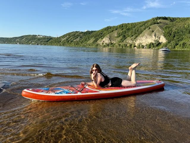 SheilaCiers with medium tits, lying on a red surfboard in shallow water, wearing black swimwear, sunglasses, and brown hair, with green hills and a boat in the background.
