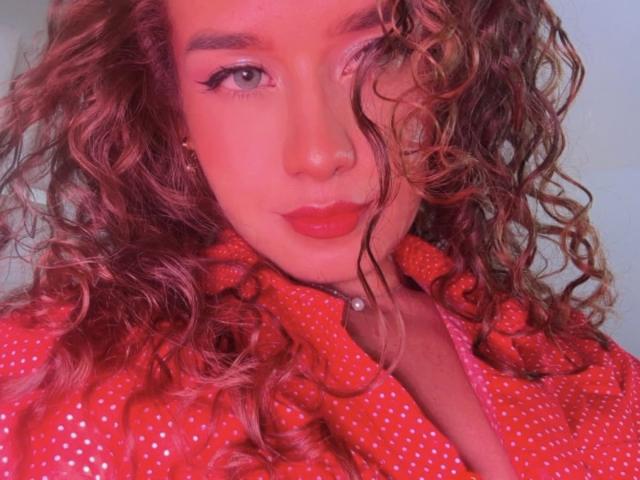 MalloryHearth with curly brown hair, red lipstick, and eyeliner, wearing a red polka dot shirt, has a pearl button on the collar, and a slightly winking expression.