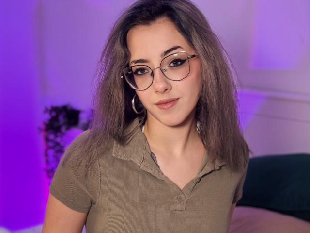 NaoimiMay with glasses, brown shirt, and gray hair, looking at the camera with a slight smile, showing her small tits and cute face, in a purple-lit room.