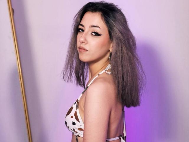 NaoimiMay with grayish-brown hair, wearing a white, brown-patterned bikini top, showing her small tits and subtle makeup with winged eyeliner, looking over her shoulder.