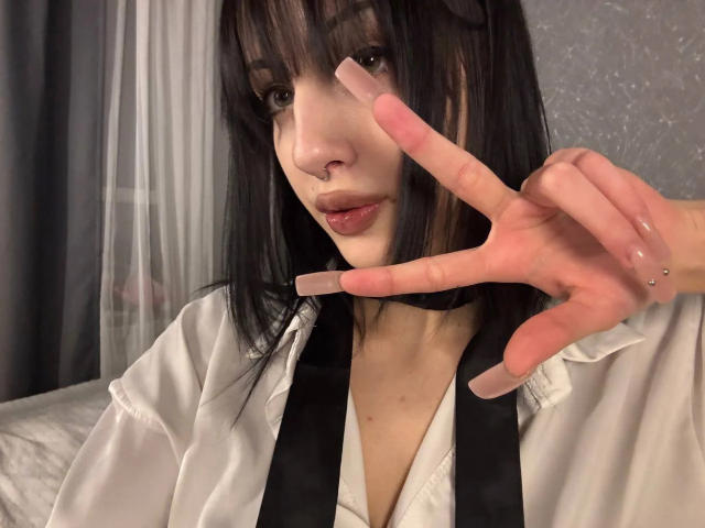 SimonneBrustkern with black hair, nose ring, and pink nail polish, flashing a peace sign with her right hand, wearing a white shirt with black suspenders.