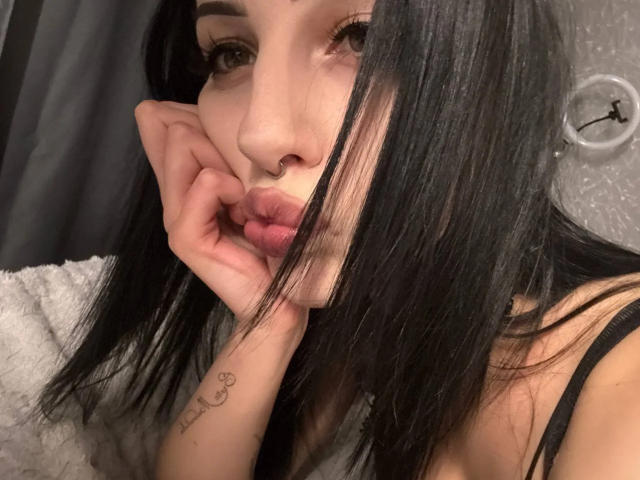 SimonneBrustkern with black hair, pouty lips, and nose ring, touching her chin with a tattoo on her wrist, wearing a black strap top. Her ass is not visible.