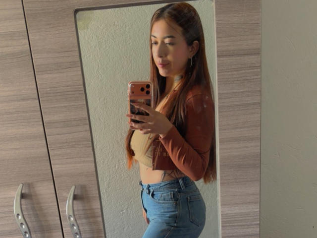 AriLynn with long brown hair, wearing a brown jacket, beige top, and blue jeans, taking a mirror selfie with an orange phone, showing her tits and ass.