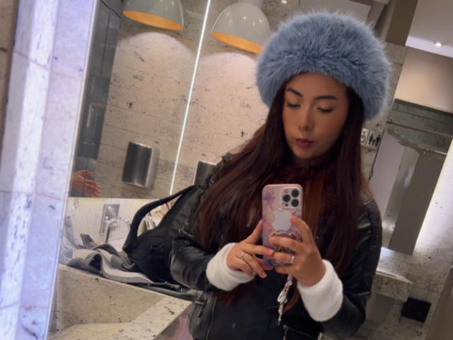 AriLynn, com cabelos castanhos longos, usando um chapéu azul de pelos, jaqueta de couro preta, tirando uma selfie no espelho em um banheiro, segurando um telefone rosa com capas brancas..