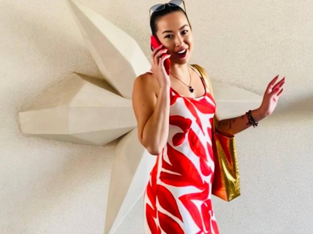 MirandaHott69 with red and white patterned dress, talking on red phone, smiling, wearing sunglasses on head, gold bag, black bracelet, and necklace, standing in front of white wall.