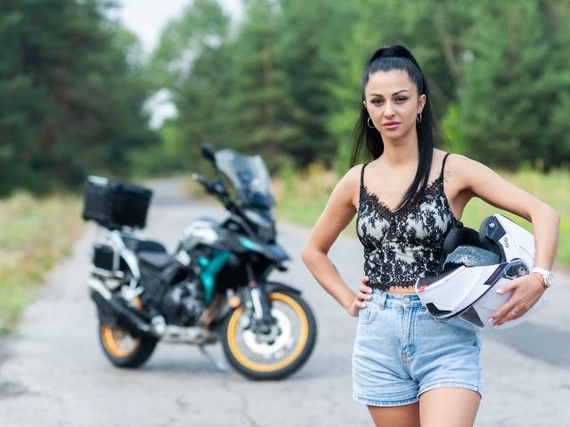 EvaClarkX with black lace crop top, blue denim shorts, holding helmet, standing in front of black and green motorcycle with a black basket on the back.