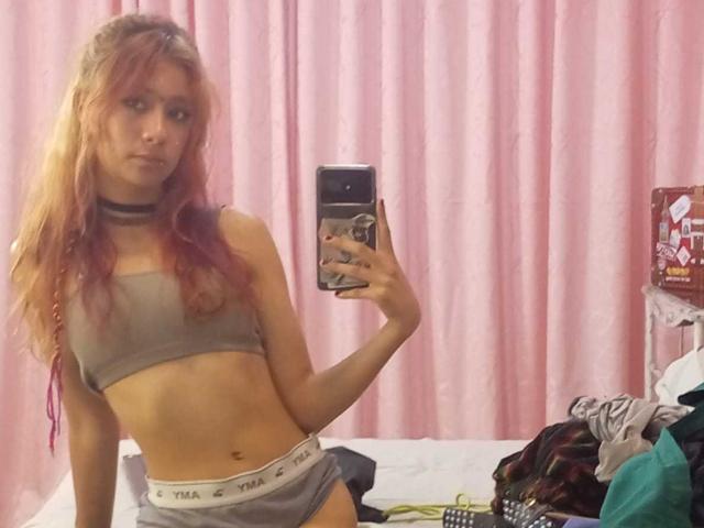 SuriEverly with long pink and blonde hair, wearing a gray sports bra and matching panties, holding a phone in a mirror selfie, showing a slim, flat-assed body.