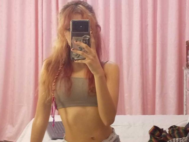 SuriEverly with long pink-blonde hair, small tits in a gray sports bra, taking a mirror selfie, holding a phone with a pop socket, in a pink-curtained room.