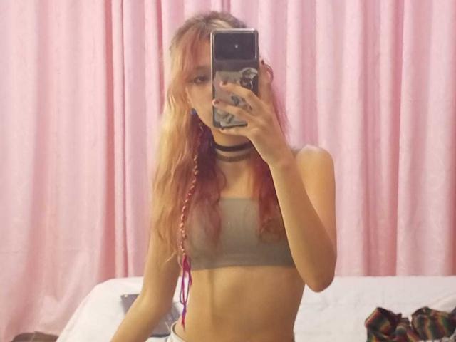 SuriEverly with pink and blonde braids, wearing a grey sports bra and black choker, taking a mirror selfie, showing a slim, flat-chested, and toned torso.