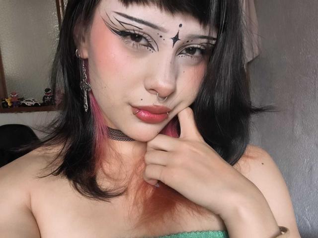 Neferetth with black hair, pink highlights, and goth makeup, holding her chin, showing off her nose ring and choker, looking at the camera with a slight smile.