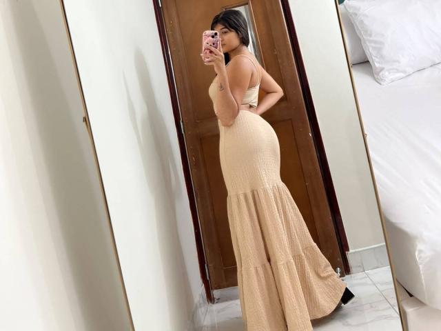 AshlyLee in a tight, beige, backless dress showing her big ass and small tits, taking a mirror selfie with a pink phone, in a bedroom.