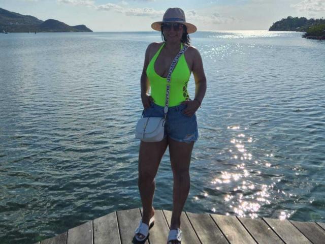 EmaFoxy met grote borsten, gekleed in een neon groen top, denim shorts, witte sandalen, een zonnehoed en een blauwe schoudertas, staat op een houten steiger aan het water..