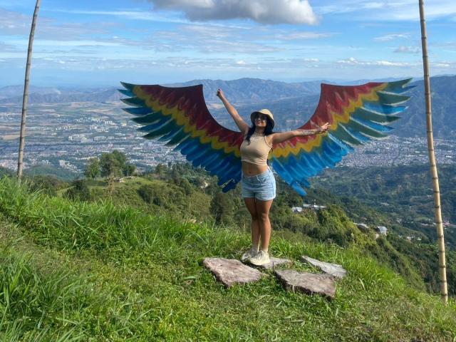 AlanaLuxe stands on grassy mountain with colorful bird wings, arms out, wearing a beige top, denim shorts, and a sun hat. Her tits are small, ass is cute.