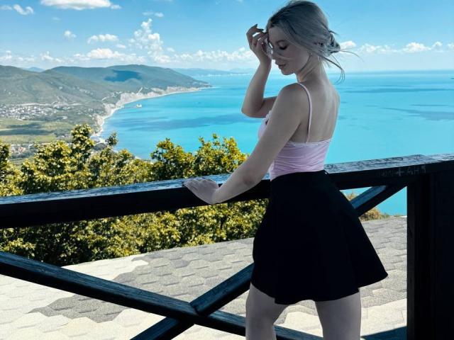 JoieGlasper with blonde hair, pink tank top, and black skirt, leaning on a wooden railing, blue ocean and green hills in the background, sunny day, holding sunglasses.