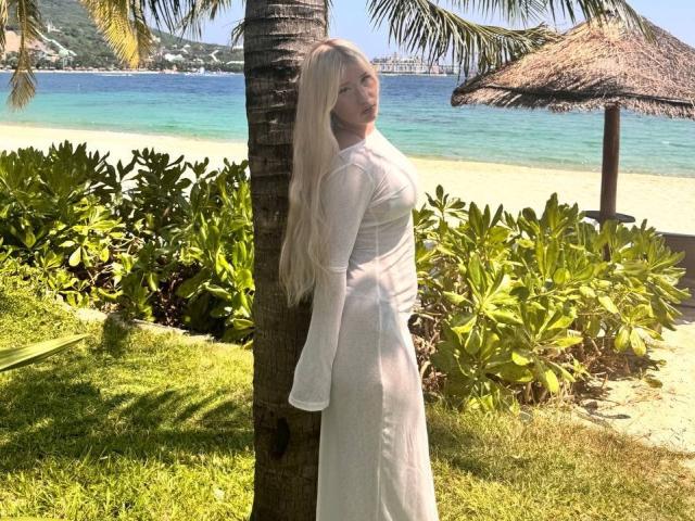 DelphiaCabos with long blonde hair, white dress, standing by a palm tree, looking back with a slight smile, her tits visible under the dress, on a sunny beach.