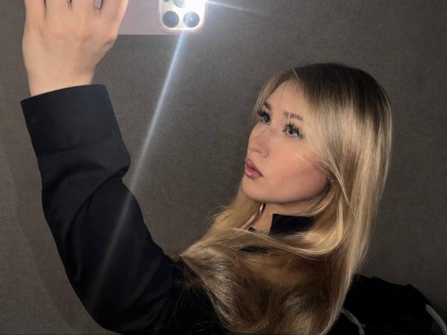 DelphiaCabos taking a selfie with a flash, showing her blonde hair, black top, and pouty lips. She's holding a pink phone, reflecting in the mirror.