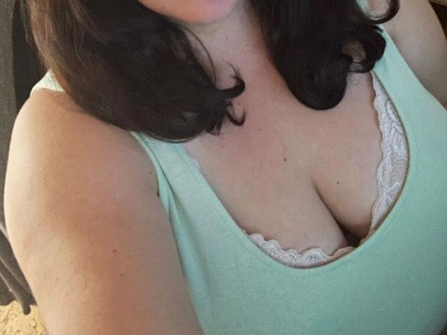 SweetGirl1998: una ragazza attraente con capelli castano scuro ondulati, che indossa una canotta verde chiaro, mostrando il suo seno generoso in un reggiseno di pizzo rosa, con pelle chiara e una leggera abbronzatura visibile..