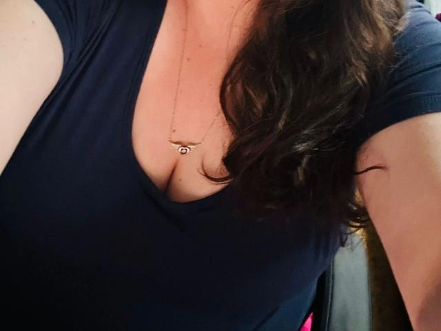 SweetGirl1998-sexy vestindo uma camisa azul escura com decote baixo, mostrando o colo e um pequeno colar com pingente, com cabelo castanho ondulado caindo sobre o ombro..