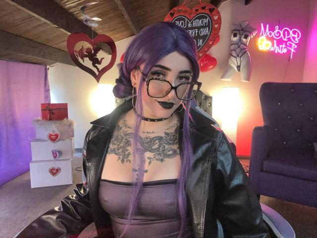 LilithDark69 with purple hair, black lipstick, glasses, black choker, leather jacket, tattooed chest, and purple tube top, showing off her big tits in a cozy room.