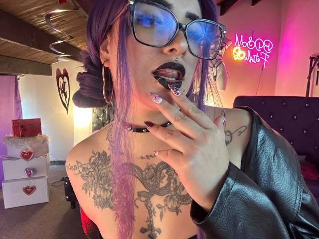 LilithDark69 met paars haar, zwarte lipstick en een bril, met haar tong uit, een zwarte leren jas, een vinger voor haar mond, tatoeages op haar borst, een choker en grote oorbellen, in een slecht verlichte kamer..
