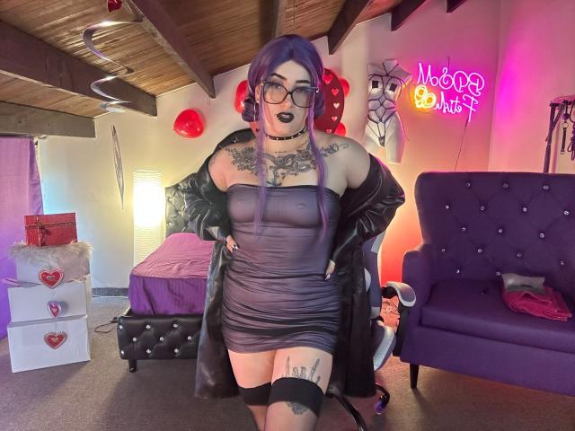 LilithDark69 tem cabelo roxo, batom preto, óculos e um vestido cinza justo. Ela está exibindo tatuagens, uma faixa preta na coxa e uma jaqueta de couro preta. Os seios estão visíveis..